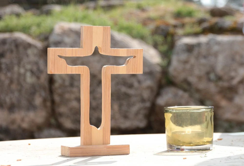 Ein Holzkreuz mit einem Ausschnitt der Silhouette Jesu steht auf einem Tisch neben einem Glas mit gelblicher Flüssigkeit. Im Hintergrund erinnern große, unscharfe Felsen und grünes Gras an die Gelassenheit, die man oft in christlichen Trauertexten findet. - Bestattungsunternehmen in Winsen, Harburg und in der Elbmarsch.