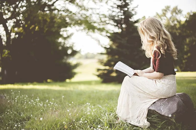 Eine Frau mit langem blondem Haar sitzt auf einem Felsen in einem grasbewachsenen Feld und liest ein Buch über Trauerbewältigung. Sie trägt ein weinrotes Oberteil und einen cremefarbenen Rock, umgeben von Bäumen und Sonnenlicht im Hintergrund. - Bestattungsunternehmen in Winsen, Harburg und in der Elbmarsch.